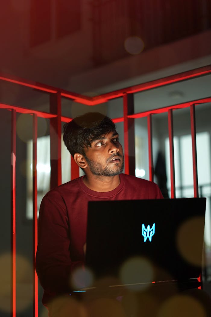 A young man sits outdoors using a laptop, illuminated by neon lights at night.
